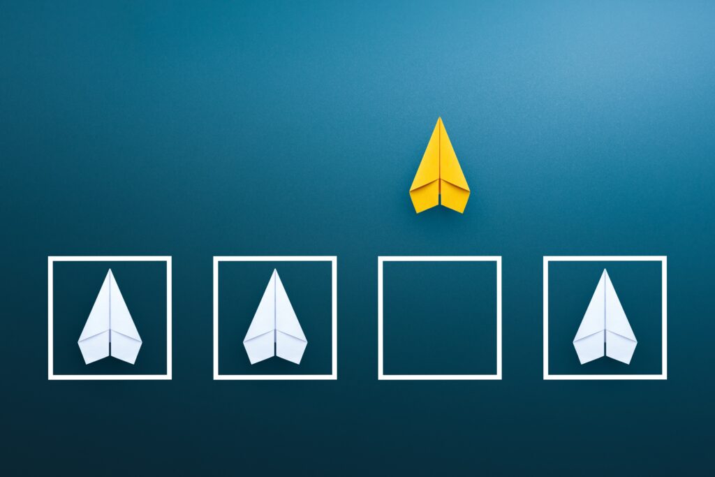 Image of a yellow paper plane outside of a box alongside three plain paper planes inside boxes to demonstrate diversity of thought